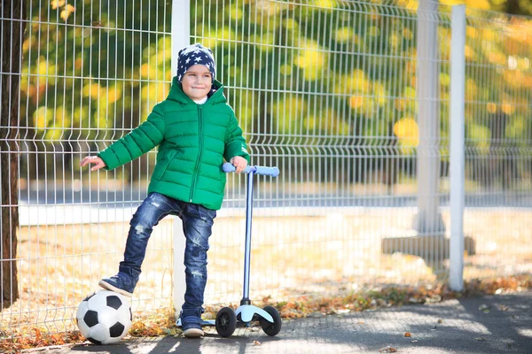 Um menino com uma bola e uma scooter está em folhas — Fotografia de Stock