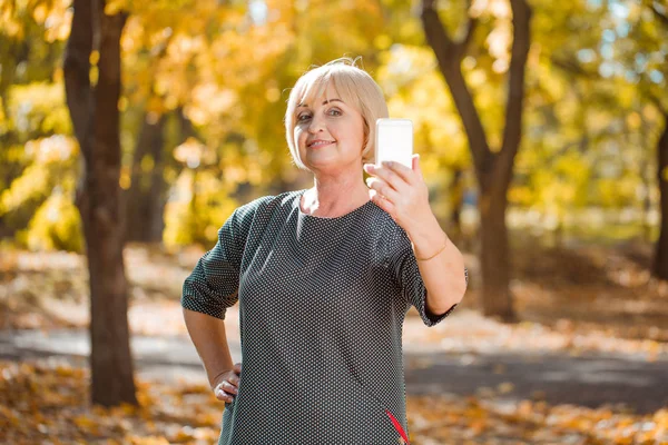 Een aantrekkelijke middelbare leeftijd vrouw lopen in een herfst park met gadgets op een onscherpe achtergrond. — Stockfoto