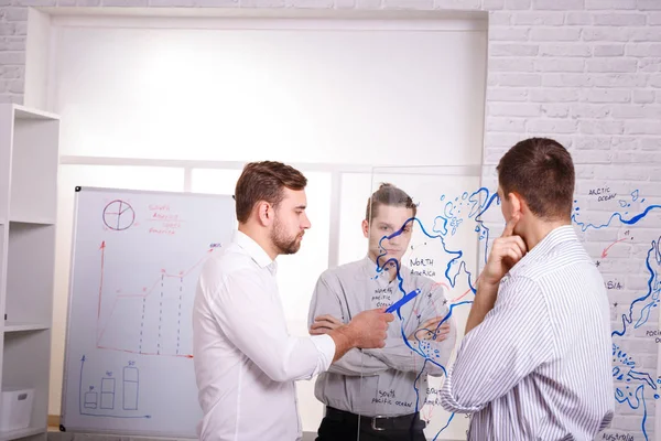 Three pensive businessmen thinking about a project and drawing a map on a transparent board in the office. Business plan concept.