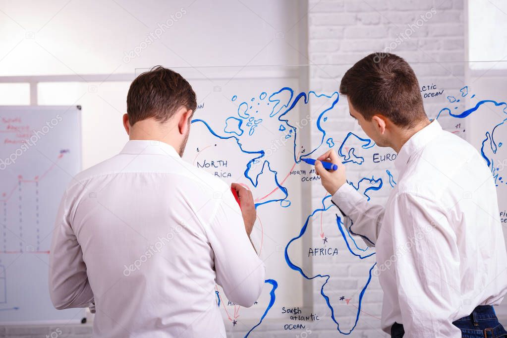 Two smart and concetrated young businessmen talking and writing on glass board in office. The concept of business presentations.