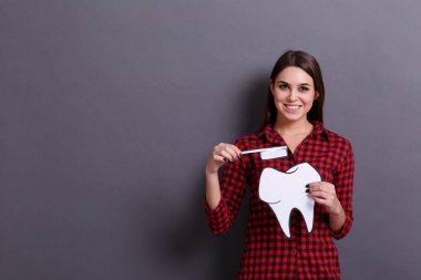 Gri bir arka plan üzerinde büyük bir diş ve diş fırçası bir kağıt kart tutan genç mutlu bir kadın. Tıp ve diş hekimi kavramı ziyaret.