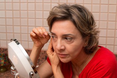 Bir kadın Ayna 'nın önünde bir çift cımbızla kaşlarını yoluyor.