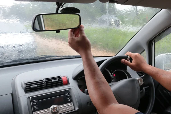 A hand of man adjust the rearview mirror of the car for better vision while raining outside of the car
