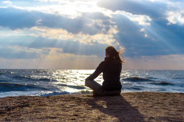 Güneşin altında denizin arka planında, kumların üzerinde oturan bir kızın silueti.