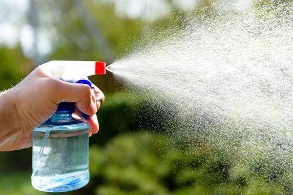 a man\'s hand sprays liquid from a spray on a background of greenery in the sun