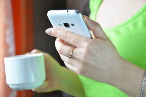 girl holds a phone in her hands and a cup of coffee. morning. against the window. morning starts on the phone. chatting news. Close up of hands woman using her cell phone in home.