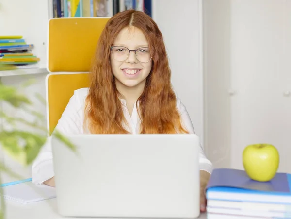 homeschooling a red-haired girl with glasses does homework at home on a laptop, student in home interior