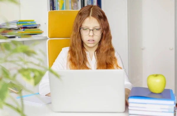 homeschooling a red-haired girl with glasses does homework at home on a laptop, student in home interior