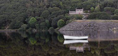 Gölde huzurlu bir manzara. Bir kayık demirli ve arkadaki ağaçların arasında bir bina var. Her ikisi de sakin suya yansıdı.