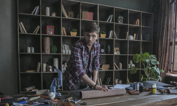 Young man leather maker unfolds roll of leather on table. Small business at home