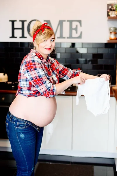 Zwangere vrouw in de keuken houden babykleding. — Stockfoto