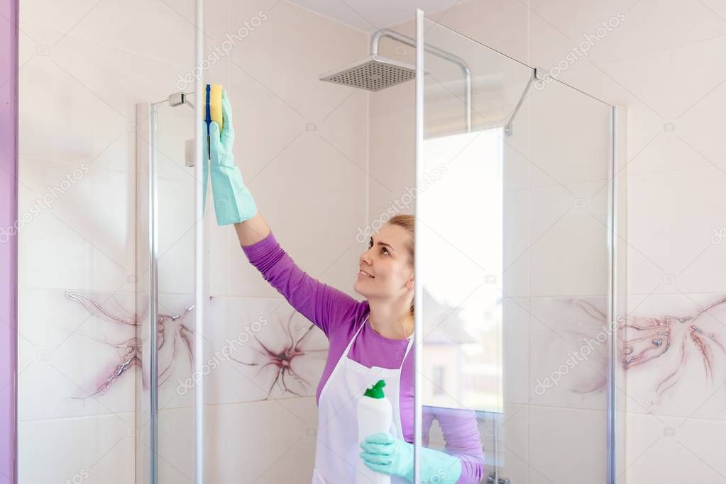 Young beautiful woman in white apron cleaning shower cabin.