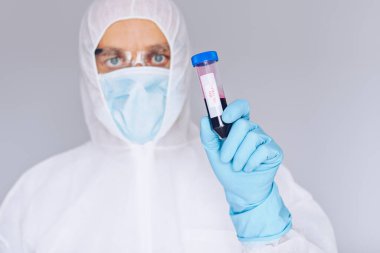 Koruyucu kıyafetli bilim adamı, elinde kanla test tüpü tutuyor.