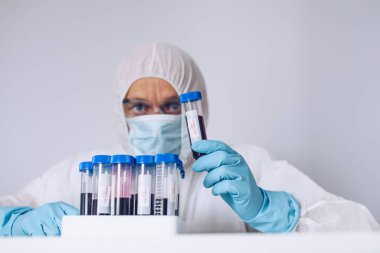 Koruyucu kıyafetli bilim adamı, elinde kanla test tüpü tutuyor.