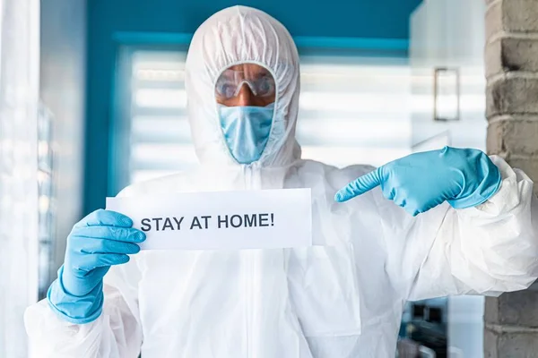 Mann in Anti-Virus-Schutzanzug und Maske hält Karte mit Botschaft zu Hause — Stockfoto