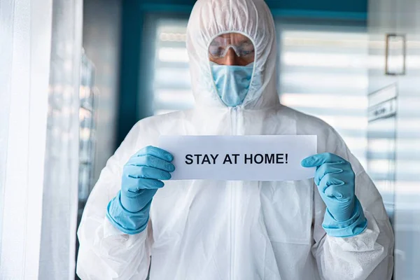 Mann in Anti-Virus-Schutzanzug und Maske hält Karte mit Botschaft zu Hause — Stockfoto