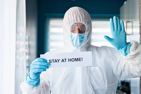Man in beschermende anti virus pak en masker holding card met bericht thuis blijven — Stockfoto