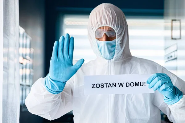 Man in protective anti virus suit and mask holding card with message stay at home in Polish language