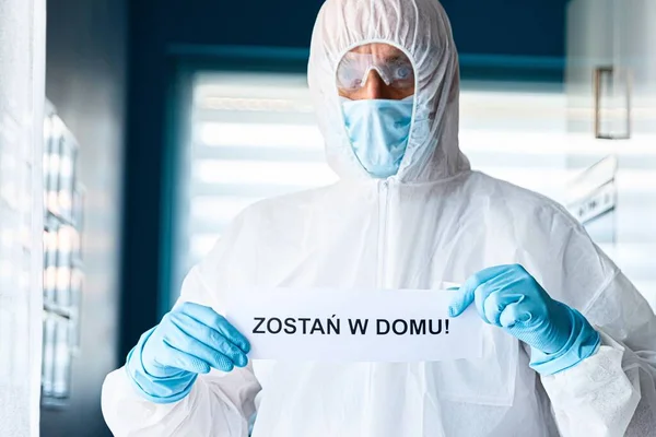Man in protective anti virus suit and mask holding card with message stay at home in Polish language