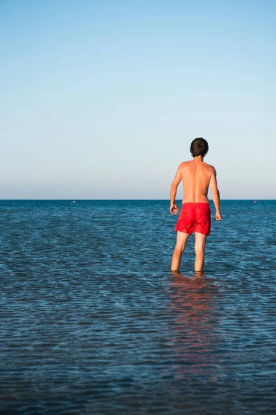瘦瘦的小伙子在海水中的红色泳装摆姿势 — 图库照片