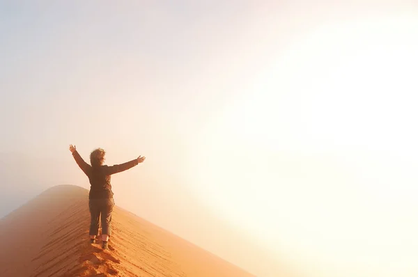 Person standing on top of dune in desert and looking at rising sun in mist with hands up, travel in Africa, Namibia — Stock Photo, Image