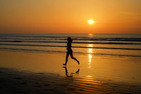 Silhouette einer Joggerin, die am Sonnenuntergang am Strand läuft, mit Reflexion, Fitness und Sportkonzept — Stockfoto