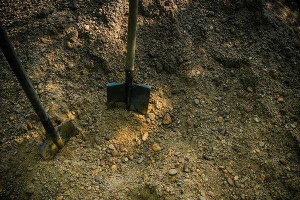 Bir kum yığını, toprak, iki küreğin yanında. İnşaat, temizlik, peyzaj. Ağır vasıfsız işçilik. Gün ışığı, üst görünüm.