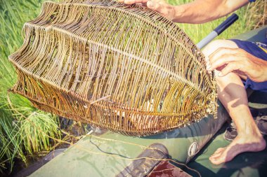 Kahverengi ev yapımı balık kapanı, eski bir balıkçı aleti. Dal kırıntıları. Arka planda yeşil bir çim var. Teknedeki balıkçı kıyıya yakın bir yerde tuzak kurar..