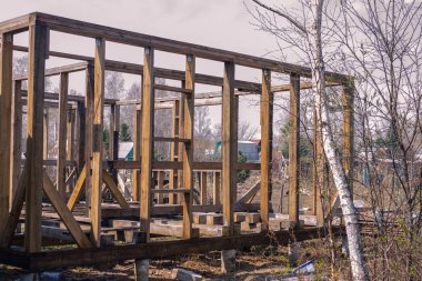 Kırsal kesimde, ahşap bir ev inşaatı. Kirişten çerçeve, ilk aşama. Güneşli bir gün, hiç kimse. Uzakta küçük ahşap evler, ağaçlar var..