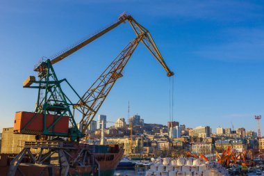 Büyük liman, liman. Gemiye kargo yükleniyor, deniz nakliyesi. Ön planda büyük bir vinç. Tepeden uzakta modern binalar var. Mavi gökyüzü, sabah.
