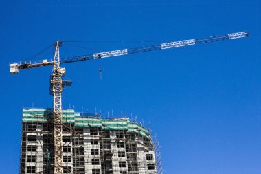 İskelede büyük bir kule vincinin yanında yüksek bir bina. Ortak inşaat, apartman dairesi. Mavi bulutsuz gökyüzü, boşluğu kopyala.