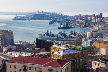 Şehir limanındaki yollarda askeri gemiler var. Donanma üssü, Vladivostok. Kıyıda birçok modern ve eski bina var. Üst manzara, güneşli sabah.