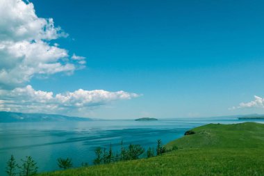 Sahilde yeşil çimenler var. Mavi su, deniz, koy, liman. Uzakta tepeler, ufuk çizgisi var. Gökyüzünde, bulutlar. Güneşli yaz günü.