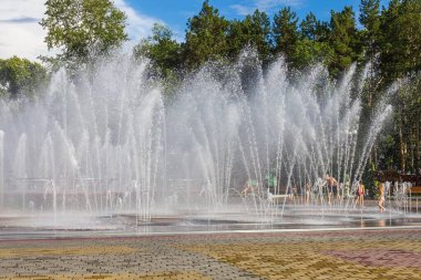 Sıcak bir yaz gününde tazelik, şehir parkında bir çeşme. Çocuklar su jetlerinin altında koşar. Mavi gökyüzü, uzaktaki büyük yeşil ağaçlar. Eğlence, huzur..