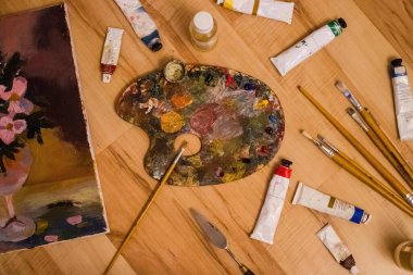Farklı boyutlarda sanat fırçaları, boyalı paletler, natürmortlu tuval, palet bıçağı. Sanatçının ustalığı, yaratıcılık, yetenek. Üst görünüm, yan aydınlatma.