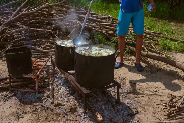 Cook cooks fish soup on the fire. Large smoked pans, camping equipment. Summer vacation in the open air. Sandy beach, green grass, firewood.