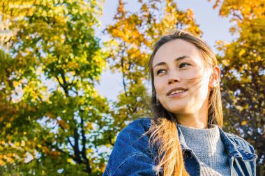 Genç bir kızın yüzü, uzun sarı saçlar, hafif bir gülümseme, yana bak. Arka planda, sarı ve yeşil yapraklar, gökyüzü. Gençlik kıyafetleri, günlük tarz..