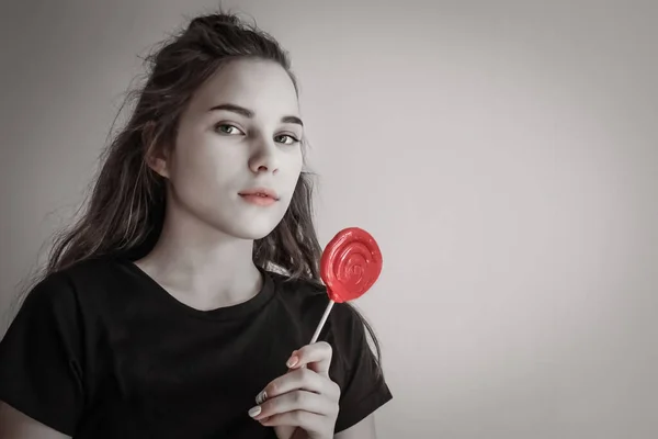 Cute girl in a black T-shirt, long hair, green eyes. In his hand is a large red lollipop. Bright moments of a carefree youth. Muted tone, vignetting.