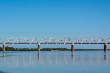 Nehir boyunca köprü. Skyline. Mavi, berrak gökyüzü. Yeşilliklerle dolu uzak kıyılarda. Pürüzsüz su yüzeyi. Köprü nehre yansıyor. Güneşli yaz günü.