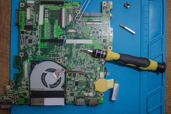 Repair laptop at home. A green board with a microcircuit, a screwdriver and a pair of tweezers. Replacement of the fuse, troubleshooting. Blue background, top view.