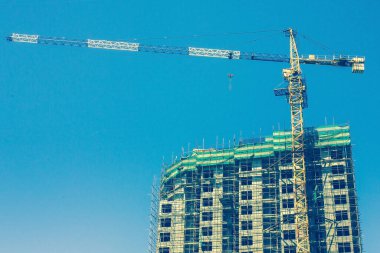 Bitmemiş bir binanın yanında büyük bir vinç. İskelenin etrafında birçok kat var. Parlak mavi gökyüzü, güneşli açık gün.