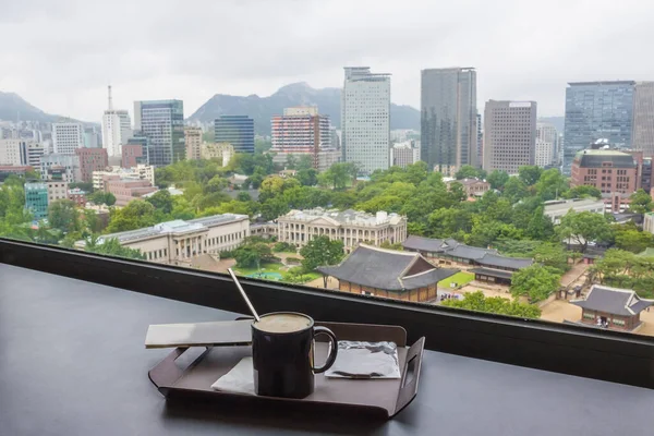 近代的なソウル ダウンタウン 窓からの眺めは 遠くの山々 手前にコーヒーのカップがあります — ストック写真