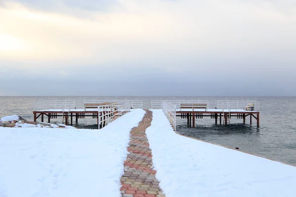 Winter beach. Blue clear sea water, snow and a wooden pier. Skyline and mountains. — Stock Photo, Image