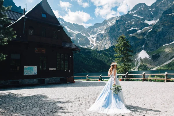 Невеста в красивом платье и венок из белых цветов держа букет цветов и эвкалипт с видом на горы и озеро — стоковое фото
