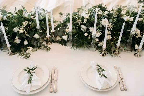 Dekorerede borde med tallerkener, knive, gafler og buket med hvide blomster og greens på midten - Stock-foto