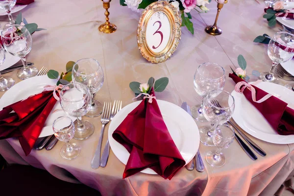 Mesas decoradas em ouro e cores borgonhas com placas, facas e garfos, velas e óculos, buquê com flores e verdes no centro — Fotografia de Stock