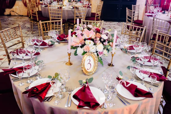 Decorated tables in gold and burgundy colors with plates, knives and forks, candles and glasses, bouquet with flowers and greens on the center