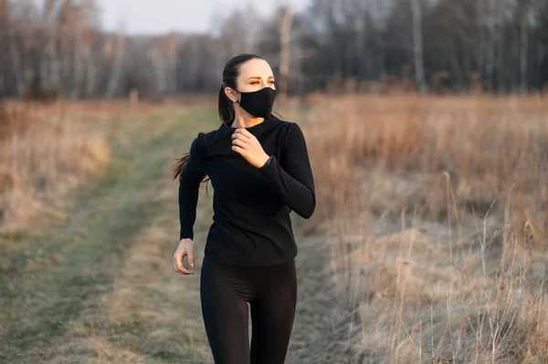 Mujer en ropa deportiva negra y máscara corre al aire libre — Foto de Stock