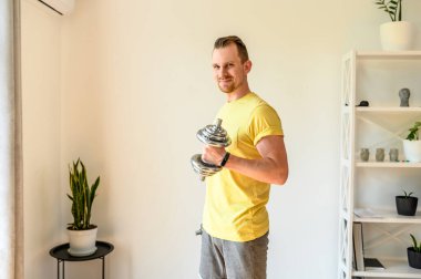 Genç bir adam evde dambıllarla antrenman yapıyor.