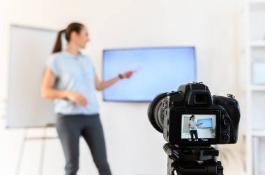 Bir kadın uzaklık çalışması için evde video dersi kaydediyormuş. Çizelge ve monitör kullanıyormuş. Ön planda tripoda kamera var. Kadın bulanık.